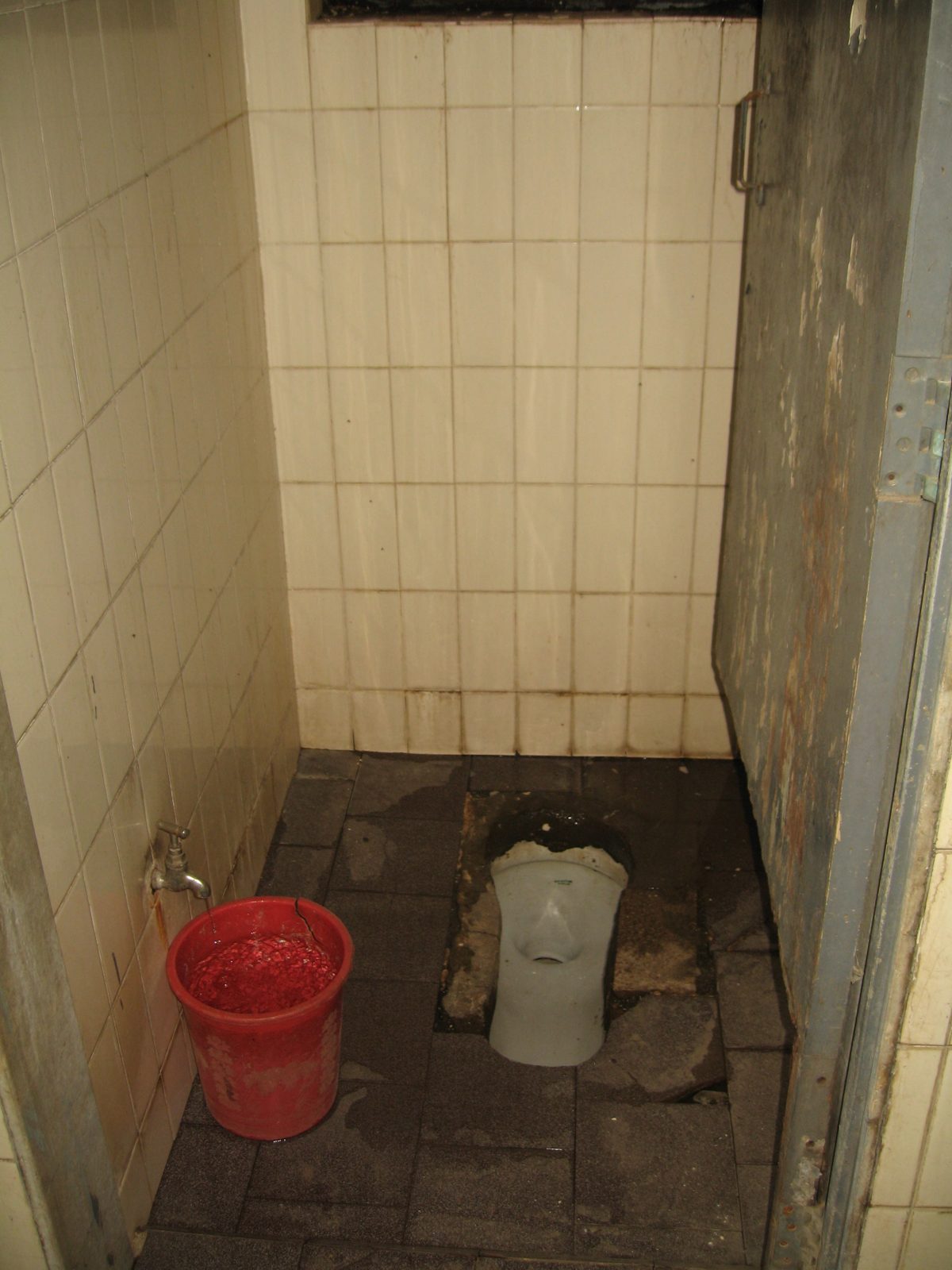 The old school bathroom: a narrow stall with a cracked, dirty squat toilet and no grab bars