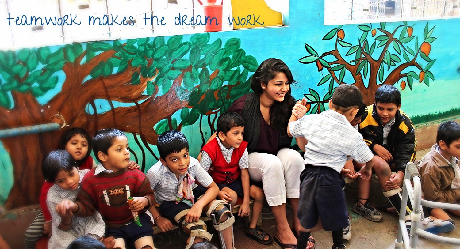 Founder Anokhee sitting and playing with a group of students in front of a painted tree mural reading "teamwork makes the dream work"